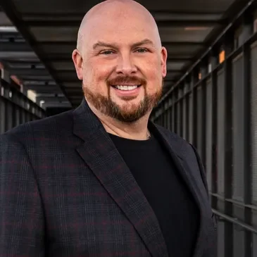 Smiling bald man in a dark blazer standing in a modern corridor.